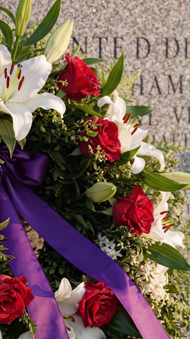 Un bellissimo omaggio floreale con una corona di rose rosse e gigli bianchi, adornata con un nastro viola e posta contro una lapide commemorativa in ricordo.