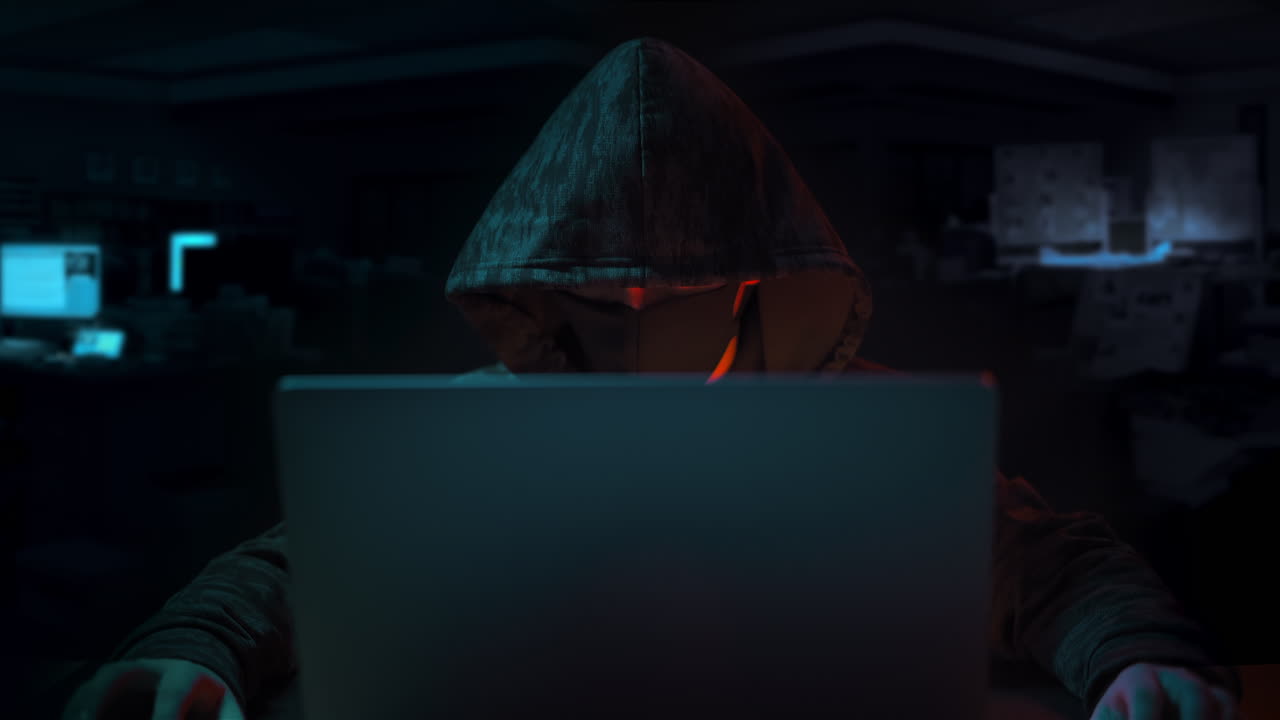 Hooded hacker with hided face sitting and watching in laptop wih hands on table in dark office room