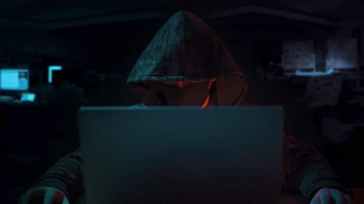 Hætteklædt hacker med skjult ansigt sidder og kigger på en bærbar computer med hænderne på bordet i et mørkt kontorlokale