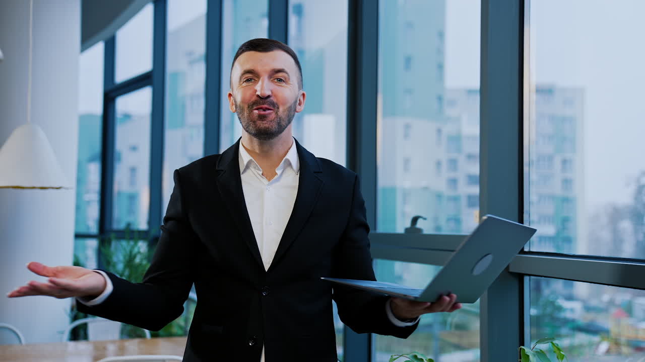 Smiling cheerful bearded man in official suit holds laptop in hands. Man speaks to the camera standing at the panoramic windows.