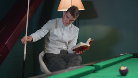 billiard player in white shirt and grey trousers sits on white chair, reading book while holding cue stick with focused expression. warm lighting enhances thoughtful and intellectual atmosphere