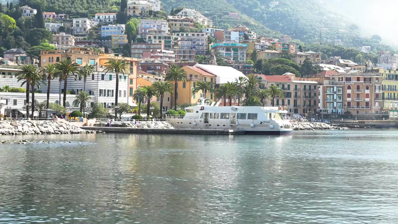 Riprese aeree di Rapallo, Italia, che mostrano l'acqua scintillante del porto, il lungomare fiancheggiato da palme e le vivaci case sulla collina sotto una luce diurna soffusa. Ampia vista panoramica senza persone visibili.