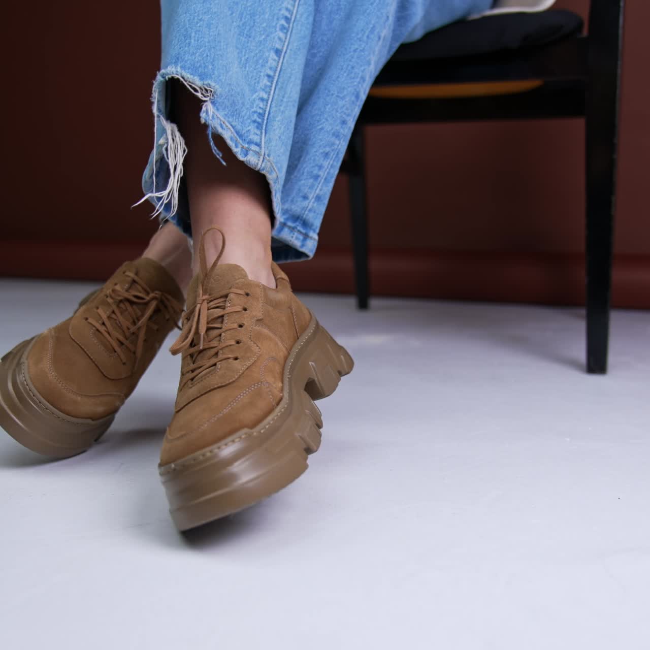 Convenient brown suede sneakers on massive soles and same color laces. Model wearing blue jeans sits on the chair posing in her shoes in front of camera. Close up