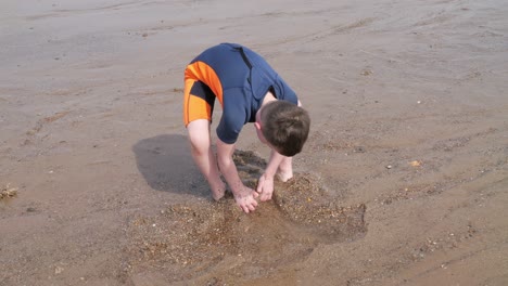 Young-boy-in-a-wetsuit-on-a-beach-digging-in-the-sand