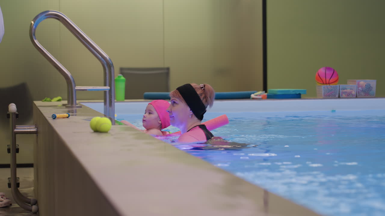 Lady taking toy from little baby while exiting pool gentle transfer bright water ripples safe supervision summer lesson smiling faces soft splashes caring guidance calm outdoor setting