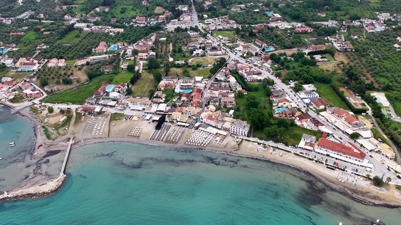 Prospettiva aerea panoramica del villaggio di Roda, Corfù, Grecia, che mostra la foce del fiume che incontra il Mar Ionio, una spiaggia sabbiosa organizzata, resort fronte mare, un ponte e infrastrutture stradali costiere.