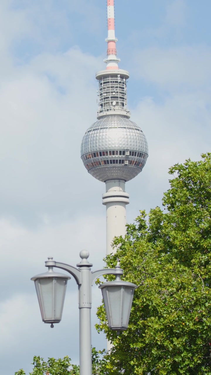 Kameraschwenk bei Tag zeigt Fernsehturm, Straßenlaternen, grüne Bäume und bewölkten Himmel in Berlin.