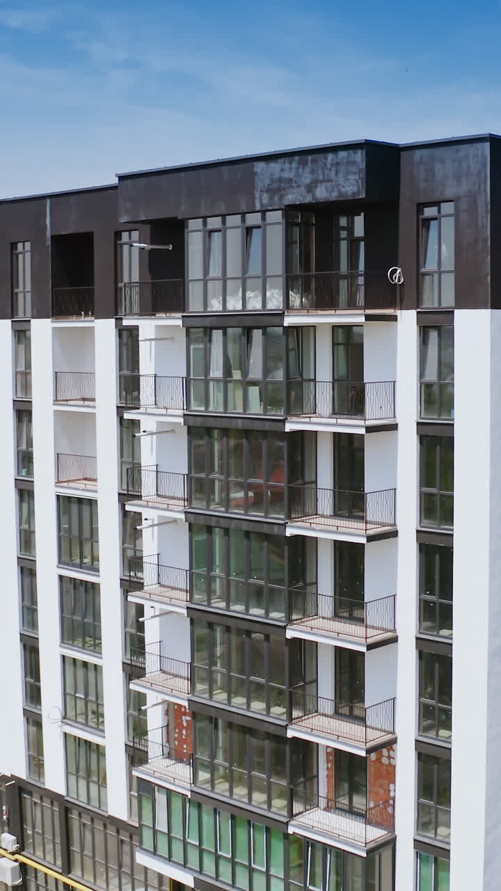 Multi-storey building in white and black colors. Exterior of a tall newly built residential apartment building with many windows and balconies. Camera moving down. Vertical video