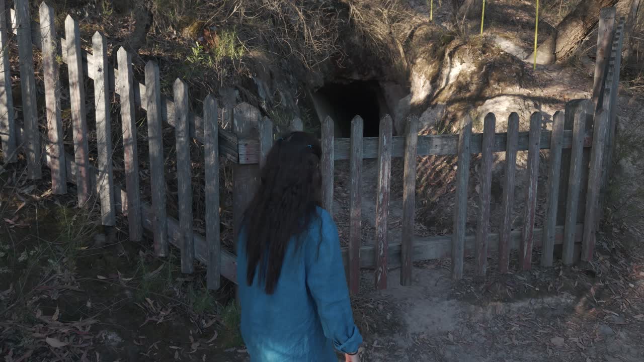 A young girl in a blue dress stands before a gated tunnel entrance surrounded by natural rocky terrain in Warrandyte. Soft daylight casts shadows, evoking curiosity and adventure