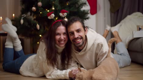 portrait of a happy couple, a guy and a brunette girl in white sweaters lie on the floor near their cream-colored cat near the christmas tree in an apartment that is decorated in the spirit of christmas