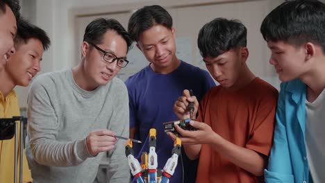 un hombre asiático con una computadora portátil, impresión 3d y una mano de cyborg enseñando a los niños a arreglar la placa de circuito en la escuela