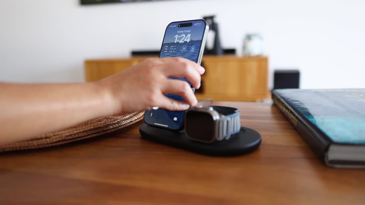 A hand places a smartphone on a wireless charger beside a smartwatch on a wooden table in a bright room