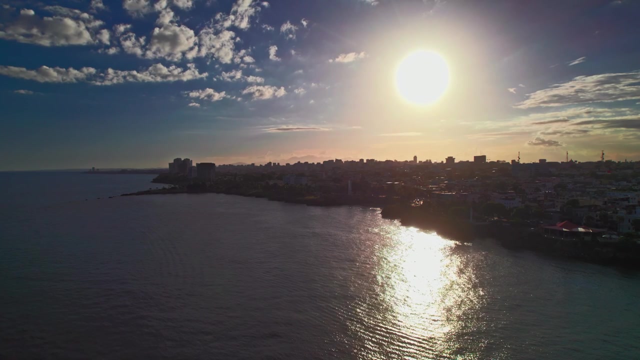 Tomada aérea estática de drone em ângulo elevado capturando a silhueta retroiluminada do horizonte da costa de Santo Domingo e o brilho intenso do sol refletindo sobre o Mar do Caribe ao pôr do sol.