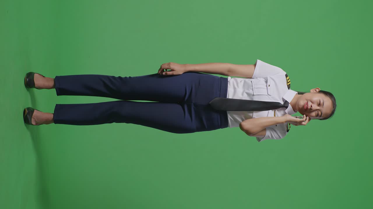 녹색 스크린 배경 스튜디오에 서 있는 동안 스마트폰으로 이야기하고 주변을 둘러보는 아시아 여성 조종사의 몸이 가득 차 있습니다.