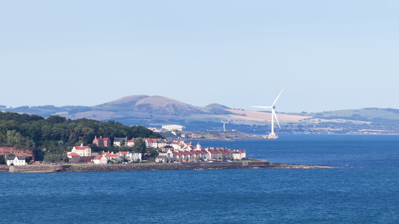 Wide shot of seaside village, wind turbine, hills, and ocean under bright daylight, static camera