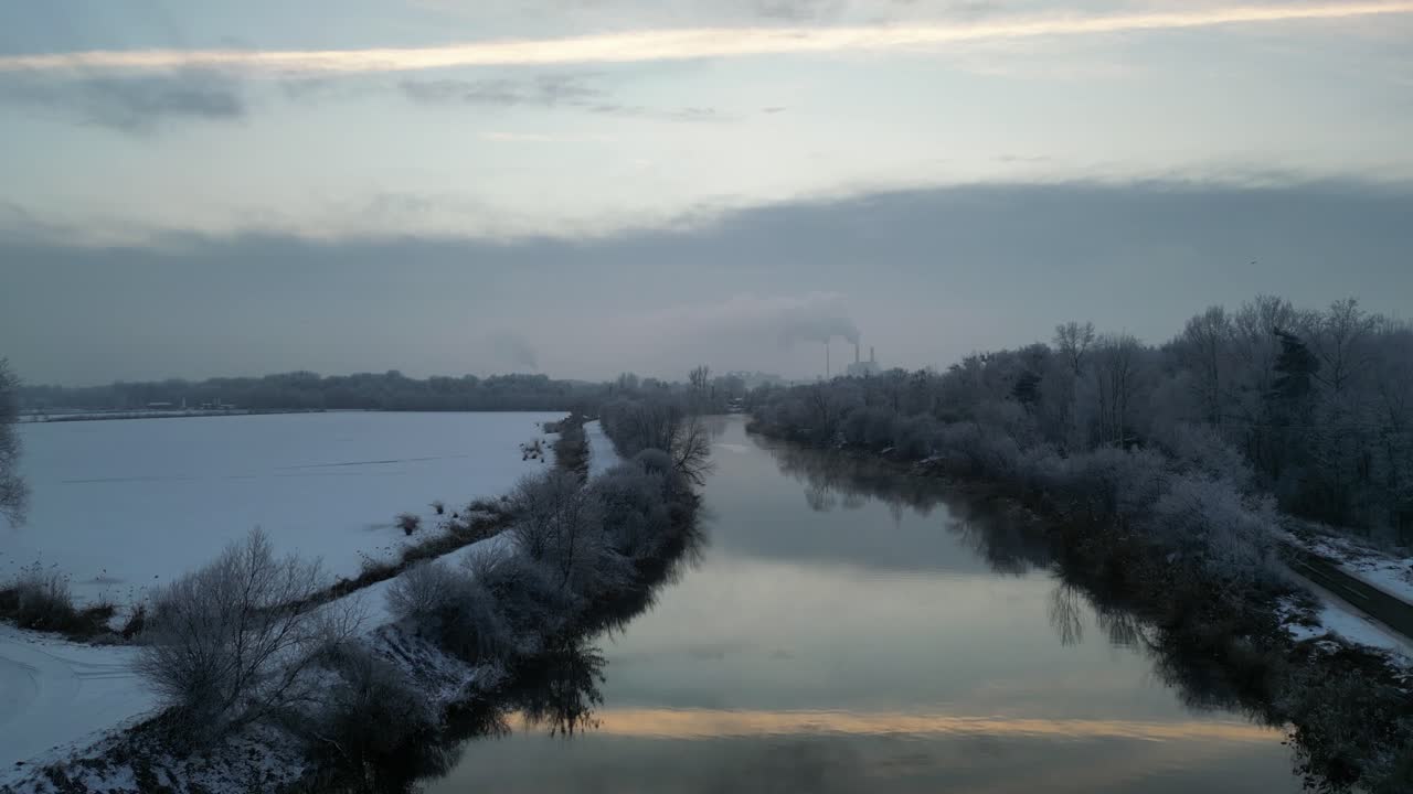 4K-luftoptagelse afslører Konins varme kanaler: Ufrosset vand frigiver damp i kraftig frost. En frossen dam ligger i nærheden, med kraftværket og røgfanen synlig i vinterhorisonten