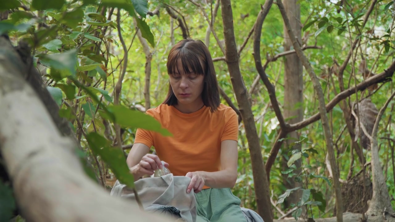 Close up of woman in orange shirt sitting among dense green foliage handling items from her bag. scene features natural soft lighting and detailed textures with visible leaves and tree