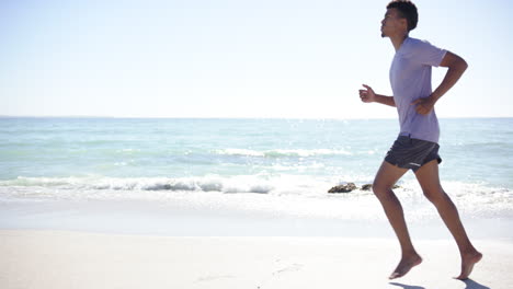 Un-Joven-Birracial-Corre-A-Lo-Largo-De-Una-Playa-Soleada-Con-Espacio-Para-Copiar,-Su-Forma-Atlética-Acentuada-Por-Casu