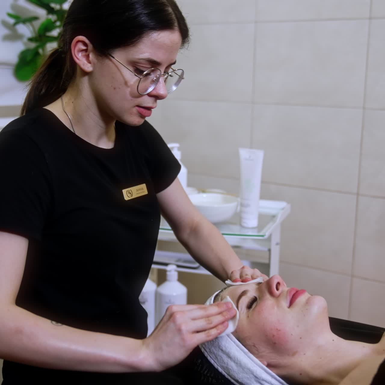 Dark-haired female in glasses uses sponges to clean the client's face. Beauty and spa procedures in the cosmetologist's cabinet
