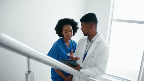 Male-Doctor-And-Female-Nurse-With-Digital-Tablet-Discussing-Patient-Notes-On-Stairs-In-Hospital