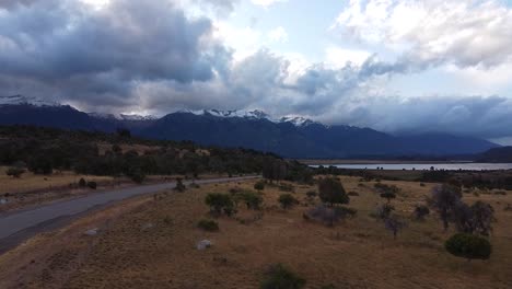 Vista-Aérea-Pasando-Por-Carretera-Con-Tráfico-De-Automóviles-Y-Majestuosos-Picos-Montañosos-Cubiertos-De-Nieve,-Capturados-En-Patagonia,-Argentina,-Sudamérica