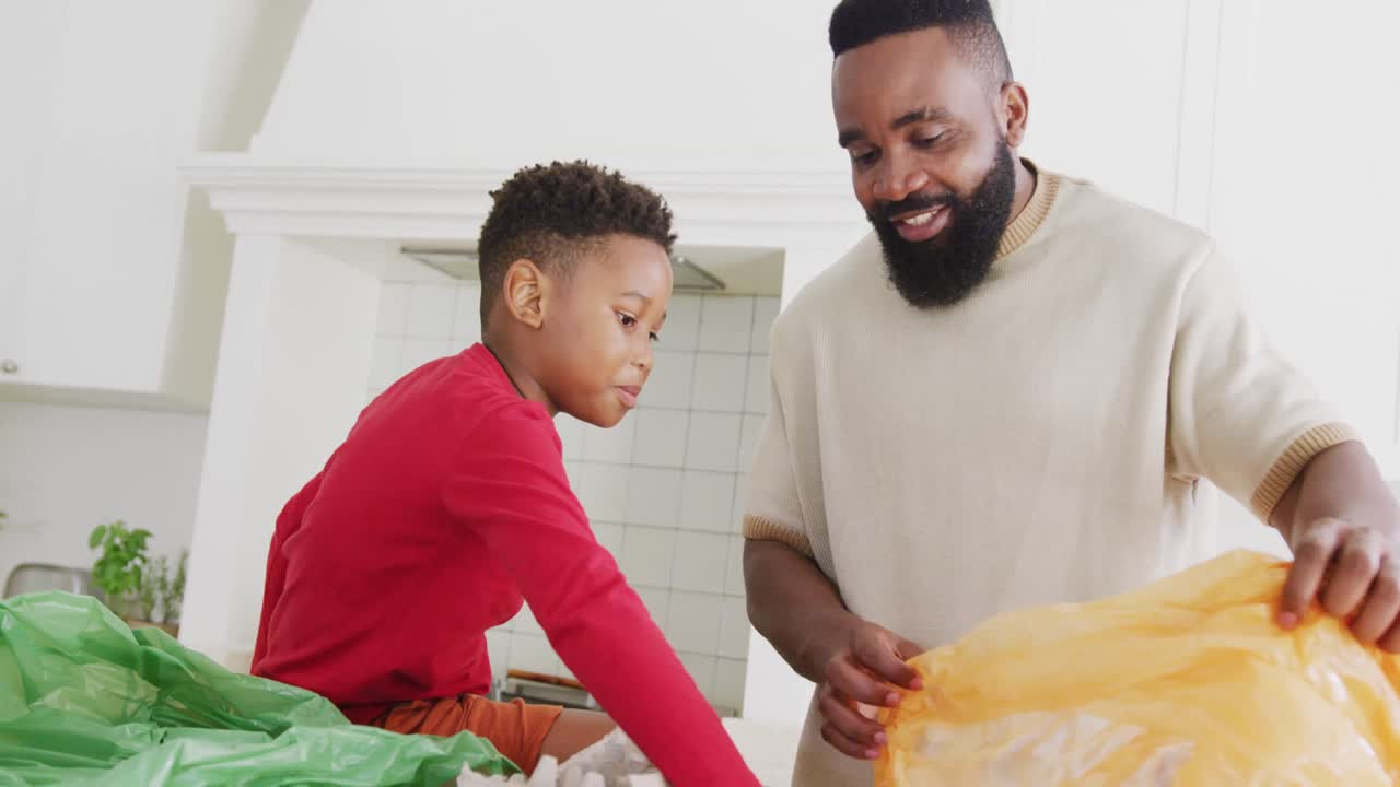 Happy african american father and son sorting waste in kitchen, in slow motion