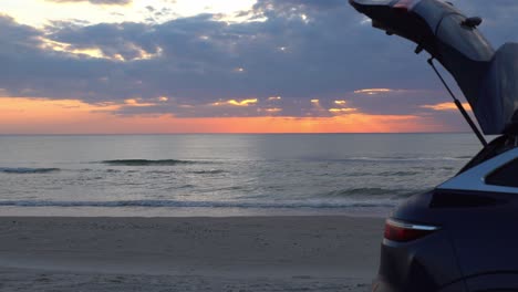 sunset over the sea with a blue hatchback car in the foreground