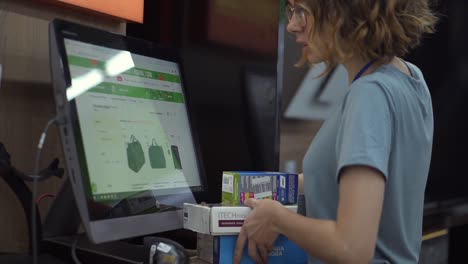 Curly-haired-slender-woman-seller-consultant-in-household-department-carry-and-scan-by-barcode-stationery-goods-in-the-boxes,-standing-in-front-the-screen.-Slow-motion