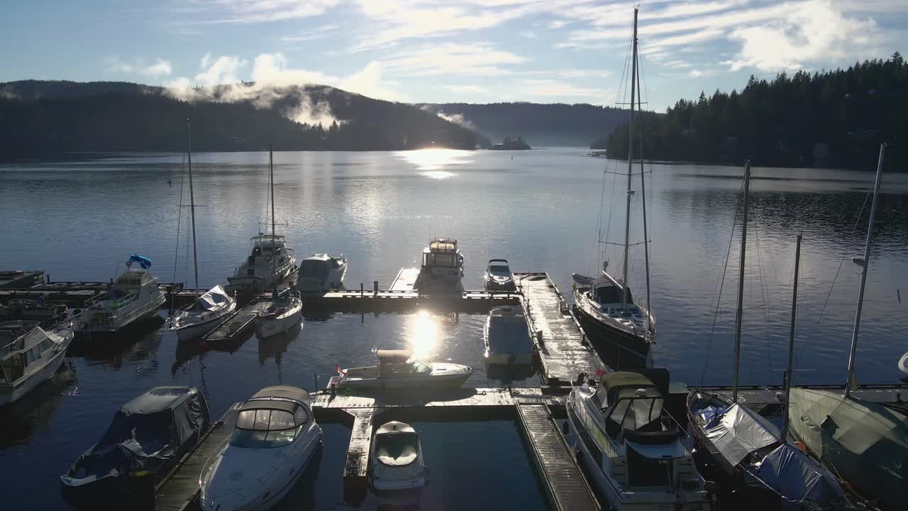 Multiple sailing yachts moored at a wooden scaffolding in a marina with the Indian Arm bay at background on a sunny day