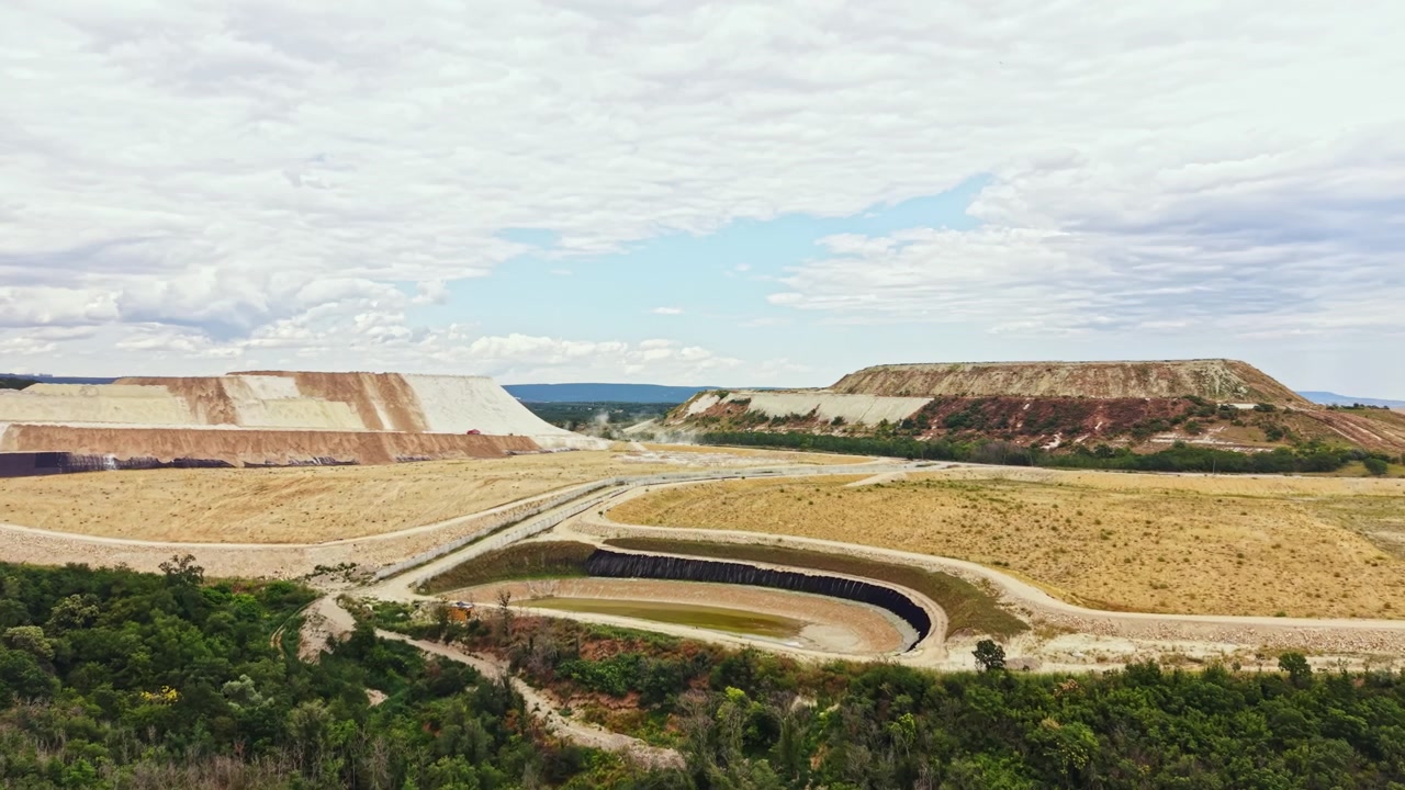 Sítio de mineração com grandes montes de terra e céu nublado ao fundo de uma área rural à tarde.