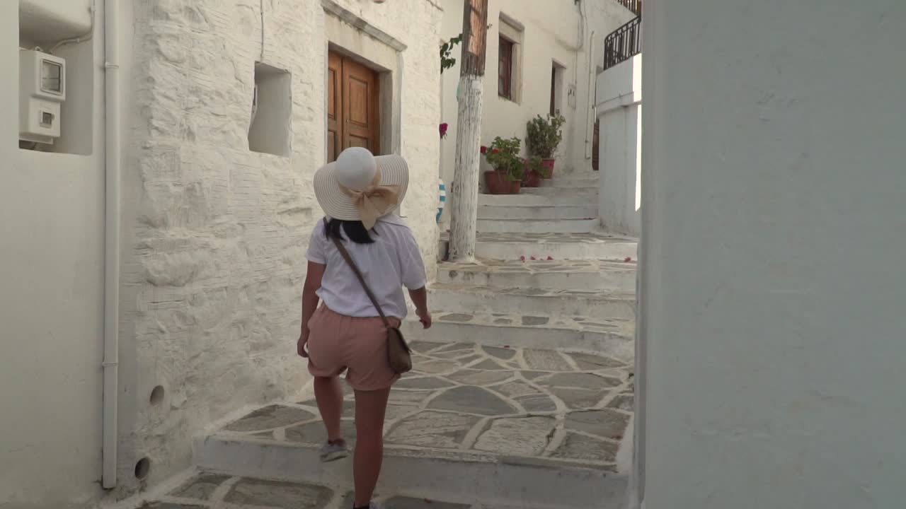 una chica vestida de senderismo con sombrero de verano caminando por un estrecho callejón griego revela