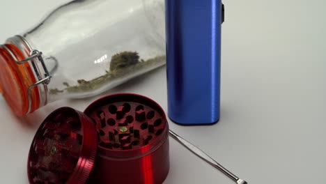 Toma-Estrecha-Panorámica-A-Través-De-Una-Cuchara-De-Metal,-Un-Molinillo,-Un-Vaporizador-Y-Un-Frasco-Transparente-Sellado-Con-Motivos-De-Marihuana