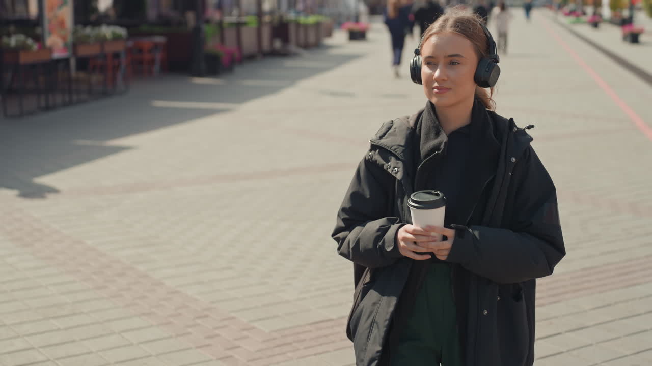 Una joven con café y auriculares hace una pausa en una plaza de la ciudad. Lleva una chaqueta negra y pantalones verdes. Este retrato espontáneo muestra una expresión relajada mientras escucha música y los viajeros pasan por su lado, con vías de tranvía.