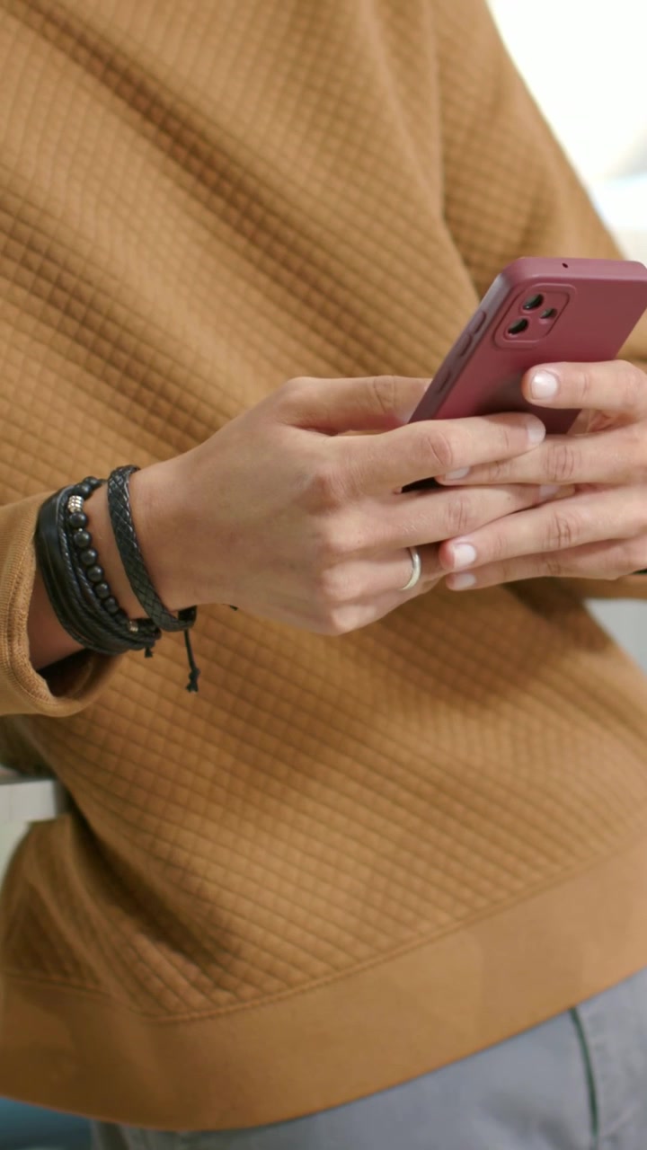 Video verticale: Uomo che tiene un telefono rosa, tocca e regola in una stanza, con pullover marrone e braccialetti.