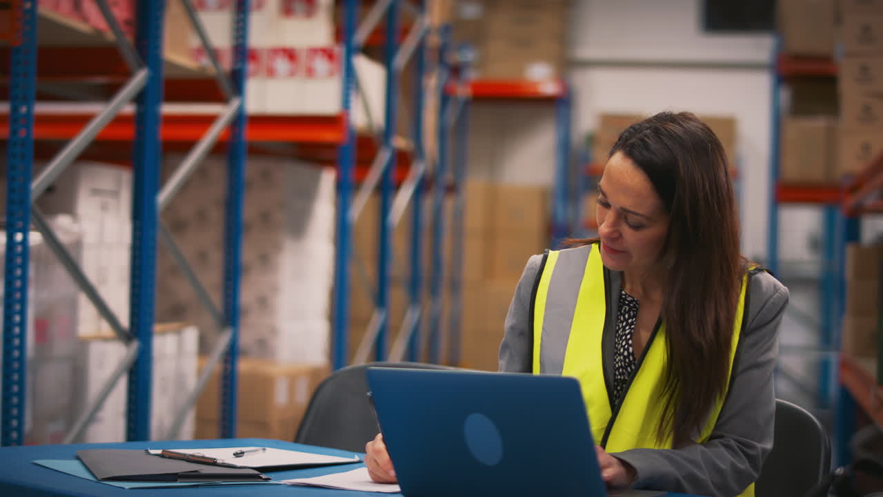 gerente femenina con chaleco de alta visibilidad sentada en el escritorio trabajando en una computadora portátil en un almacén ocupado