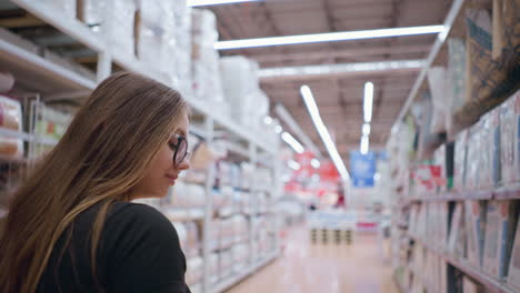 back view of woman in black top adjusting hair while looking at products on store shelf, blurred retail environment with bright lighting, stocked textile goods, and modern supermarket interior