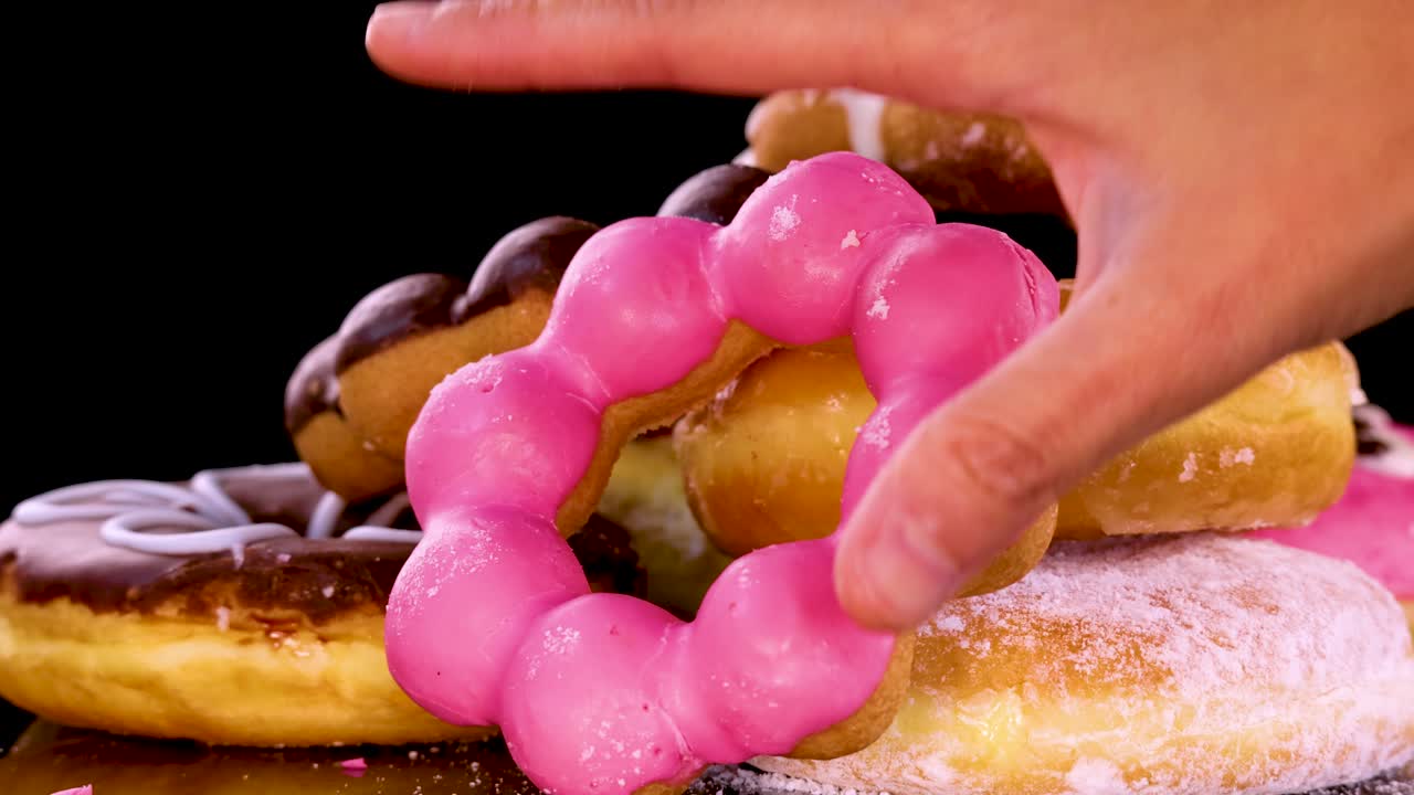 A hand reaches into a stack of assorted donuts, picking up a pink frosted ring donut. Bright studio lighting and black background highlight the vibrant colors