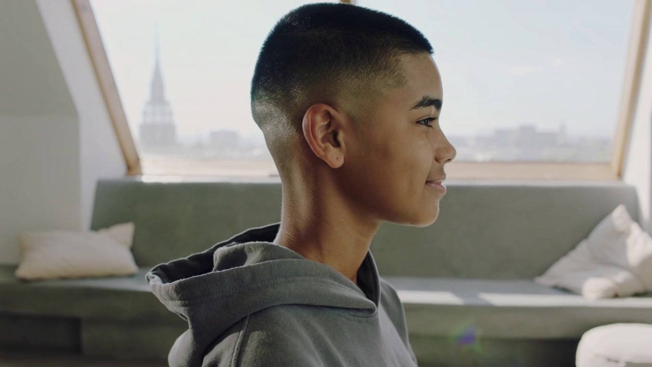 Turning teen in gray hoodie pivoting head to right profile in loft, camera opening, skylight flare