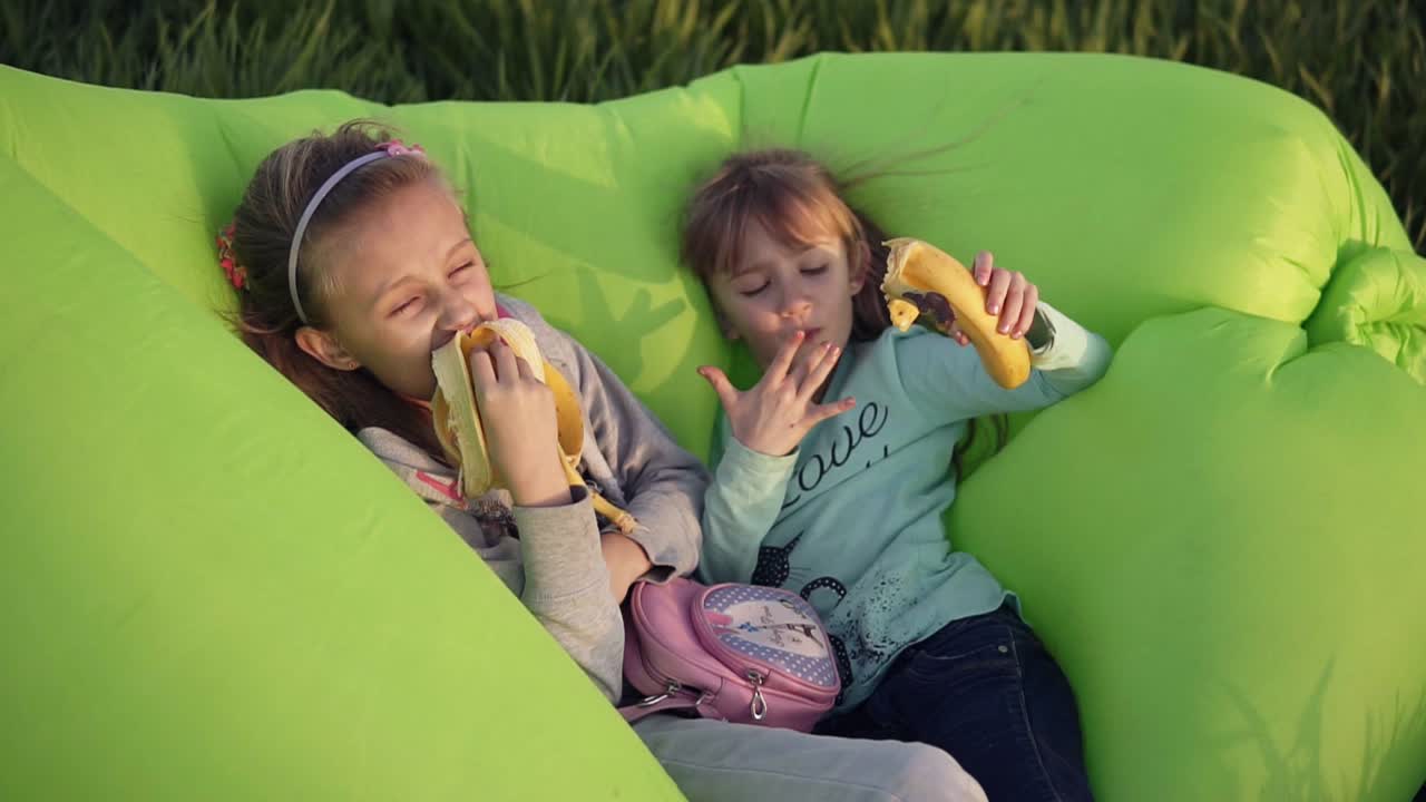 dos hermanas pequeñas acostadas en un sofá verde inflable al aire libre. descansando, comiendo plátanos. la pequeña lamiendo dedos. sabroso. día soleado. hierba verde
