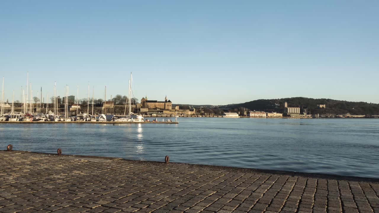 Ludzie w porcie w Oslo o zachodzie słońca, z Twierdzą Akershus w tle, szerokokątny film poklatkowy.