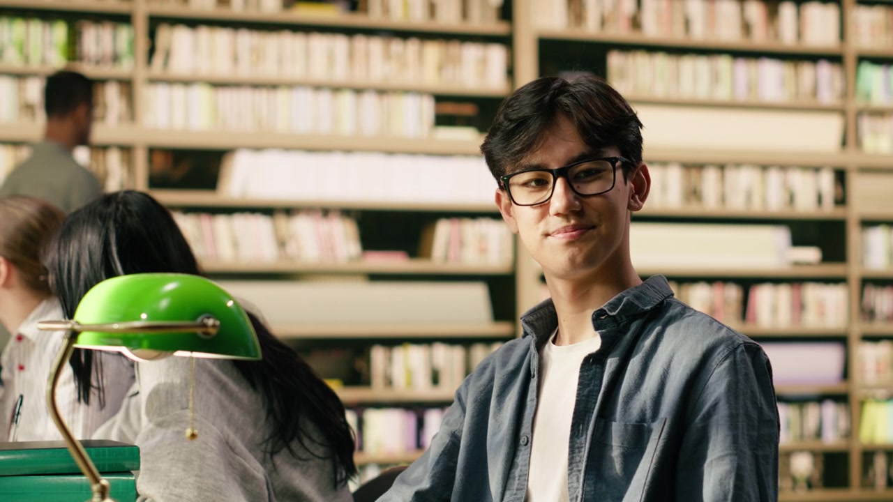 Retrato de joven con gafas leyendo un libro abierto en una mesa de biblioteca de madera.