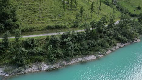 Pintoresca-Escena-Al-Aire-Libre-De-Un-Sereno-Cuerpo-De-Agua,-Un-Lago-Ubicado-Junto-A-Una-Carretera,-Un-Paisaje-Enriquecido-Por-Una-Exuberante-Colina-Verde-Adornada-Con-árboles,-Una-Roca-Visible-En-La-Hierba-Cerca-De-La-Carretera,-Un-Atractivo-Estético