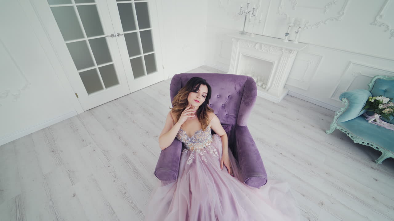 Luxury young woman with perfect makeup and beautiful dress in a chair in studio. Attractive bride in wedding dress is posing to camera while sitting in armchair in stylish room.