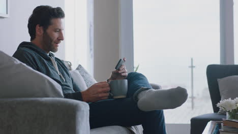 joven usando un teléfono inteligente navegando en línea leyendo mensajes de redes sociales disfrutando de un estilo de vida cómodo bebiendo café relajándose en el sofá en casa