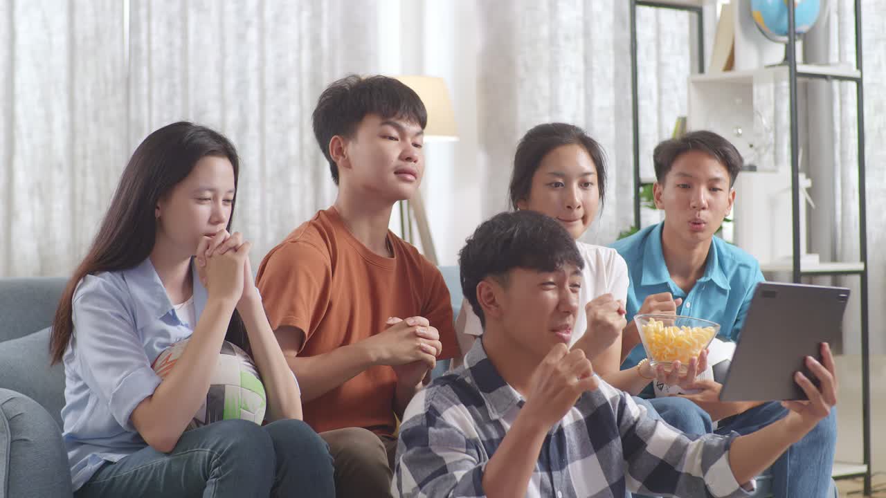 아시아 십대들이 태블릿에서 축구 경기를 응원하고 지켜보고 집에서 실망합니다.