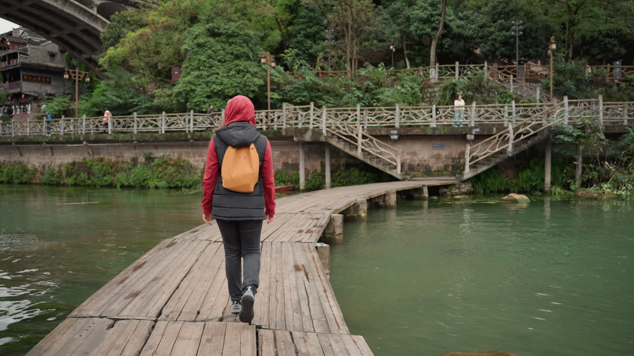 Backpacker solo berjalan di jembatan kayu di atas sungai hijau menuju jembatan gantung pedesaan, jaket merah dan ransel kuning, latar belakang hutan yang tenang, cahaya alami yang lembut, langkah santai, pantulan ambien.