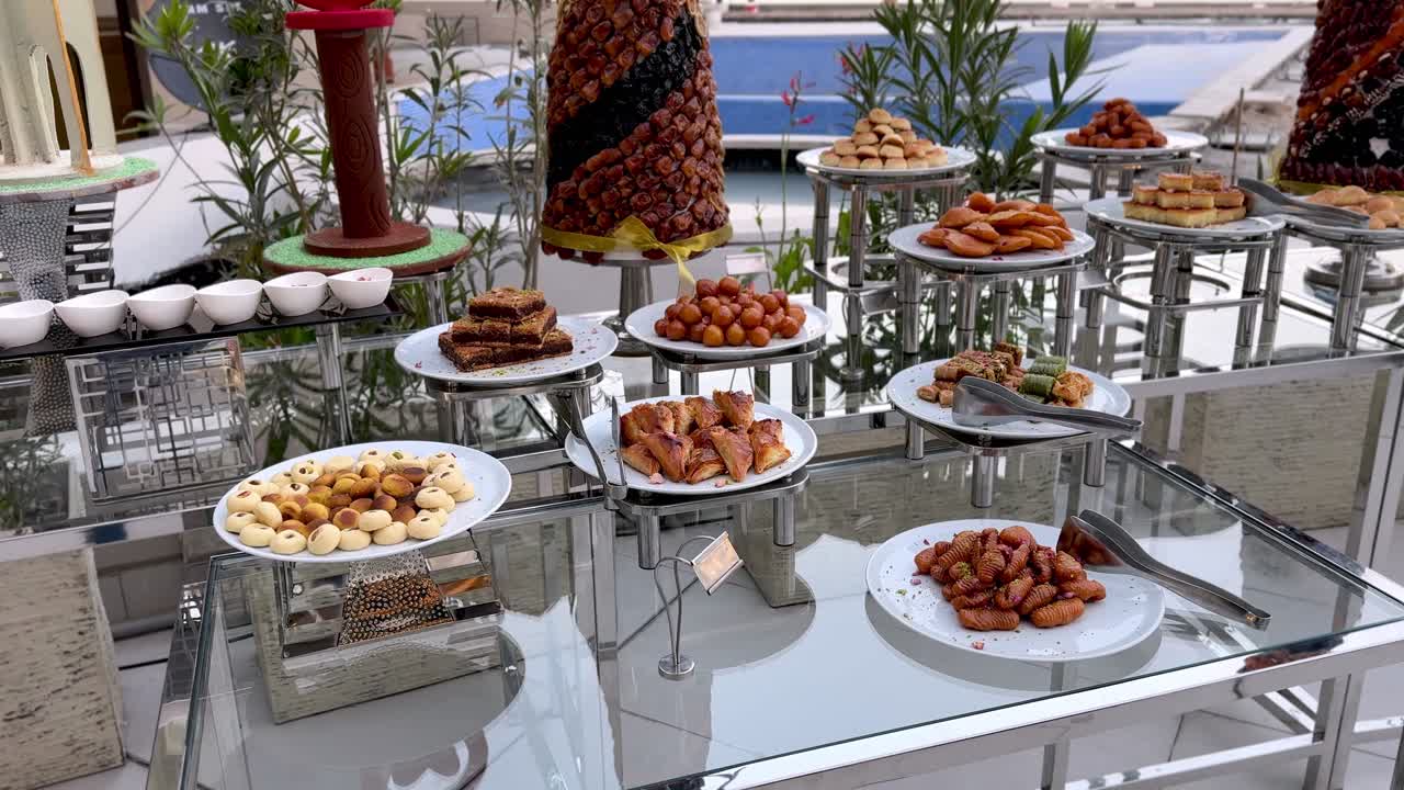 Elegante buffet di dolci a bordo piscina con dolci tradizionali arabi, baklava, pasticcini ripieni, gnocchi sciroppati e torri di datteri decorative su tavoli di vetro a un evento di catering di lusso