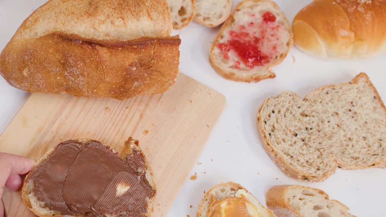 Mano che spalma crema al cioccolato su pane a lievitazione naturale su un tagliere di legno, su sfondo bianco.