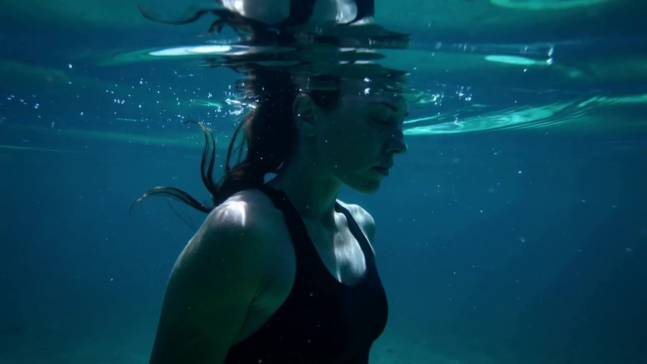 Donna atletica in costume da bagno nero che galleggia in posizione verticale sulla superficie dell'acqua, con bolle che salgono.