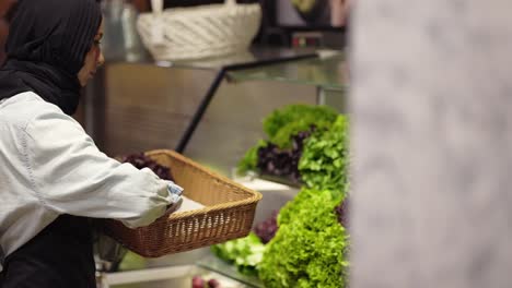 woman in scarf refill the fresh greens on the shelf at the supermarket, side view